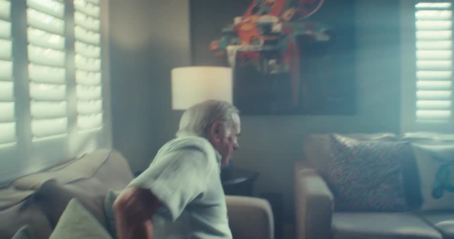 An elderly man turns around smiling and sits down on the couch in a living with sunlight streaming in through the window