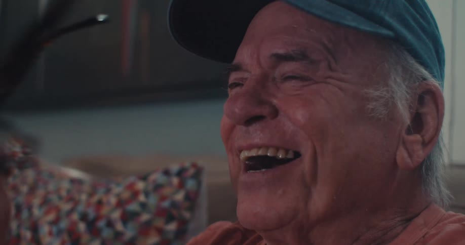 An elderly man laughs with joy and happy tears on a couch indoors