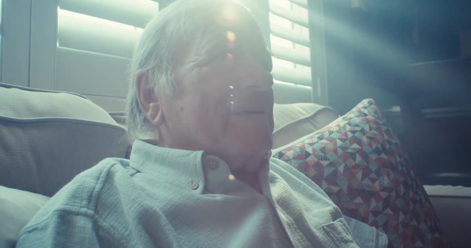 An elderly man sits on a couch smiling next to a window at midday
