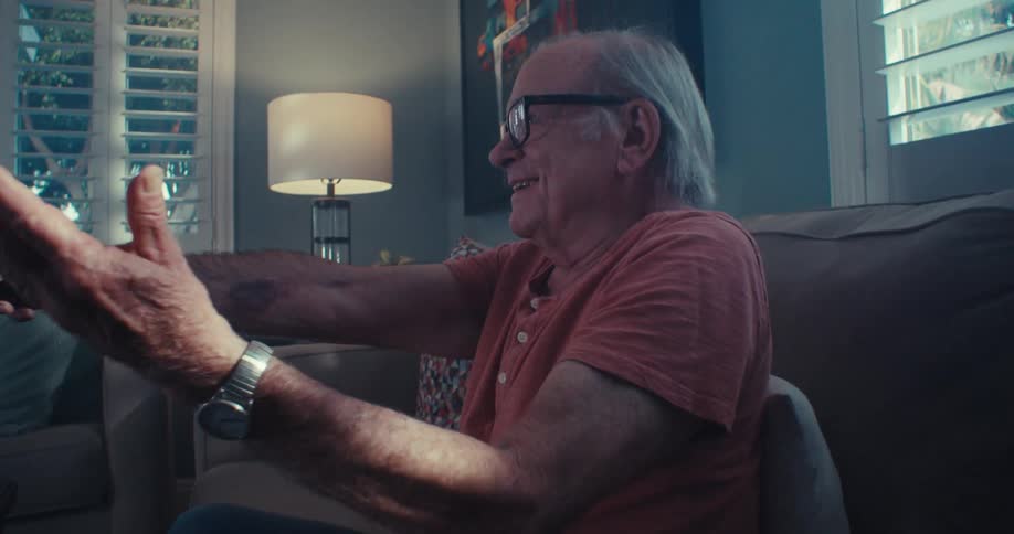 An elderly man smiles and uses a remote control to operate an electronic appliance while sitting in a living room