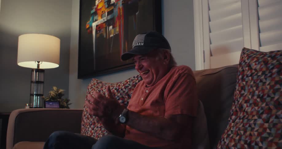 An elderly man wearing a blue hat that says veteran excitedly claps and smiles as he looks at his tablet while sitting on the couch