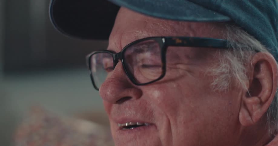 An elderly man wearing a veteran cap and glasses smiling and laughing while watching television