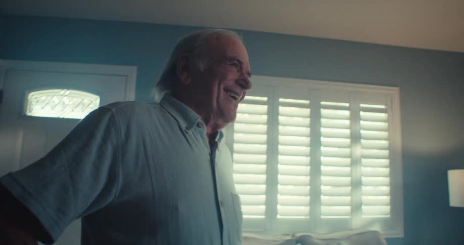 An elderly male dances the jig in his living room as sunlight filters through the window blinds in the background