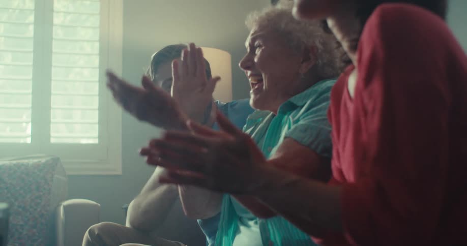 An elderly woman sits on a couch between a couple indoors filled with excitement as they cheer and clap their hands while watching a tv show