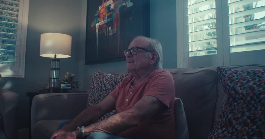 Elderly man laughs and watches tv while sitting on a couch in a living room