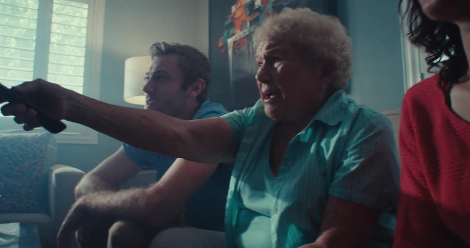 An elderly woman sitting between a man and a woman on a couch holds a remote control and points it at the tv while complaining