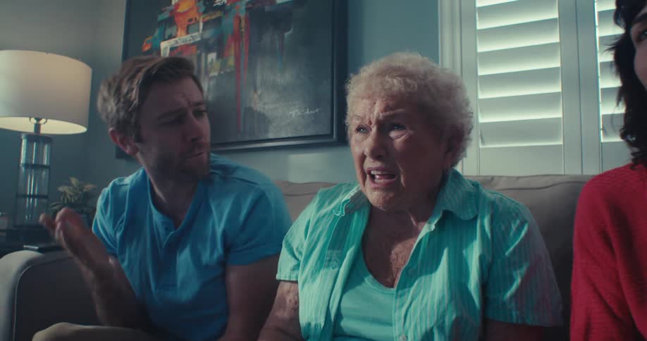 A family of three a man an elderly woman and another woman sit on a couch passionately watching and reacting to something off screen