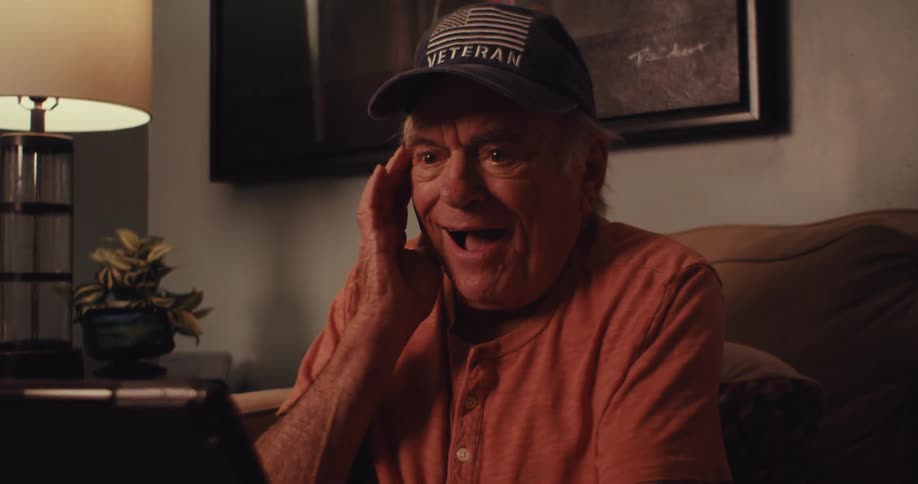 An elderly man wearing a cap is looking at a tablet that is in his hand and he is making emotions of surprises then starting to smile
