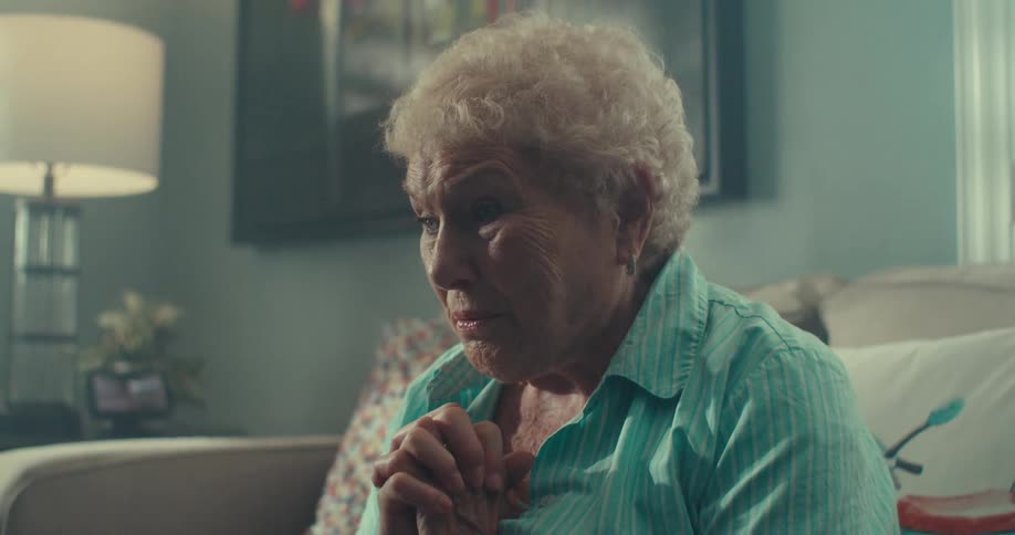 An elderly woman with white hair is looking forward in nervous tones and she folds her hands