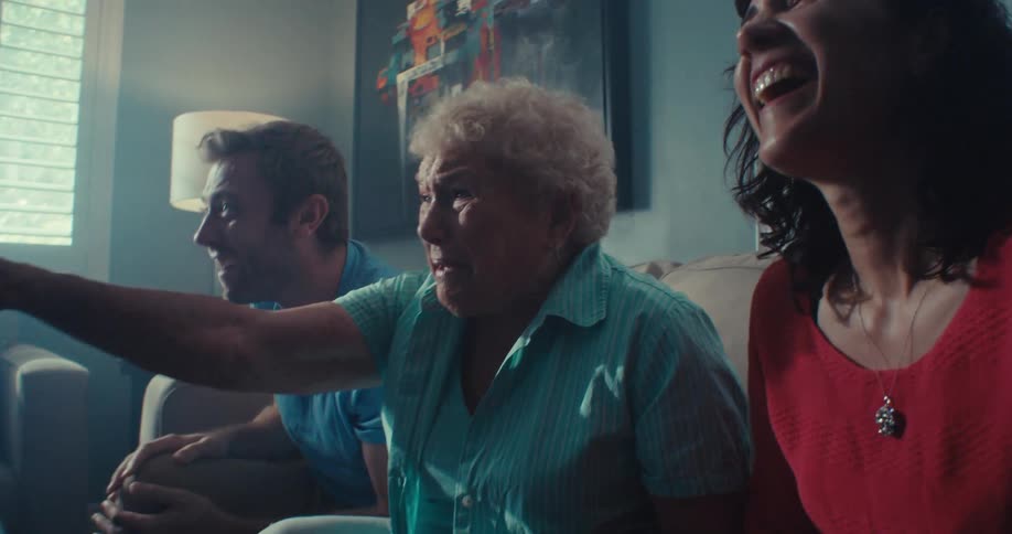 An elderly woman sits on a couch between a couple indoors filled with excitement as they cheer while watching a tv show