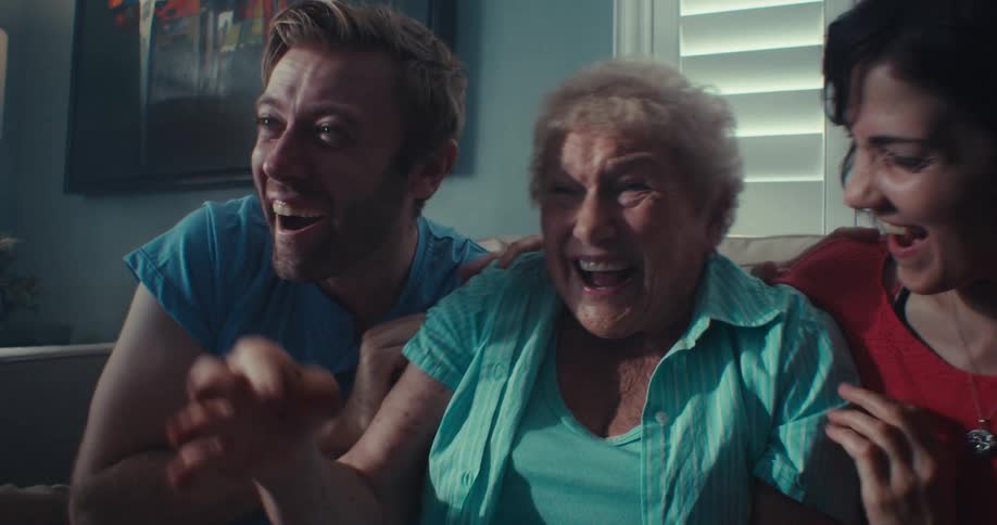 Two young people and an elderly woman are sitting on a sofa and are anxiously looking at something in front of them and then they start to celebrate and the elderly woman claps her hands