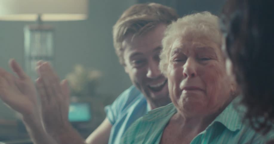 An elderly woman sits on a couch between a couple indoors filled with excitement as they cheer while watching a tv show