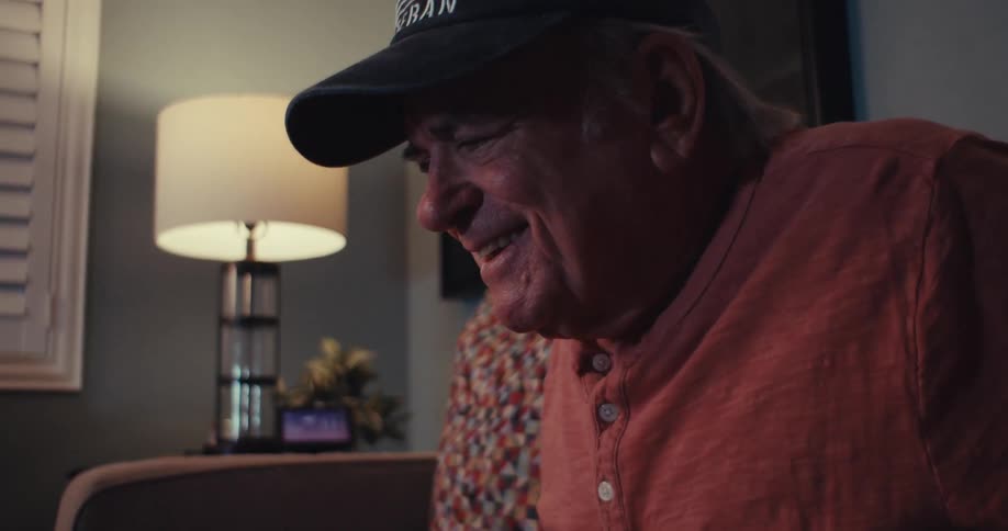 Elderly man in cap sitting smiling in a living room at night