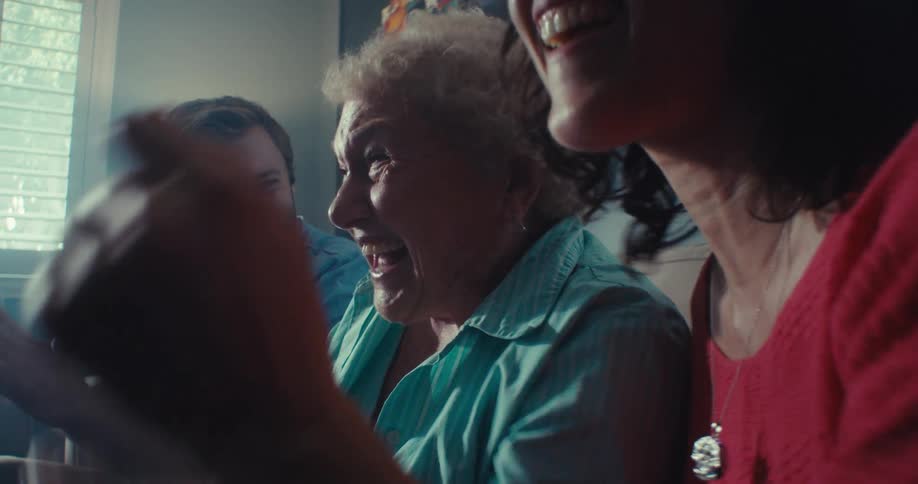 An elderly woman cheers excitedly on a couch with two smiling adults in a cozy sunlit room