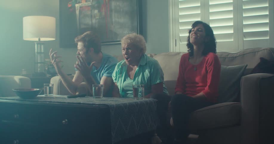 Two young people and a lady are sitting on a sofa in an indoor environment and are looking forward to a tv in nervous tones