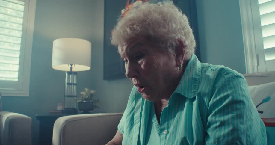 An elderly woman intently uses a laptop while sitting on a couch in a cozy living room