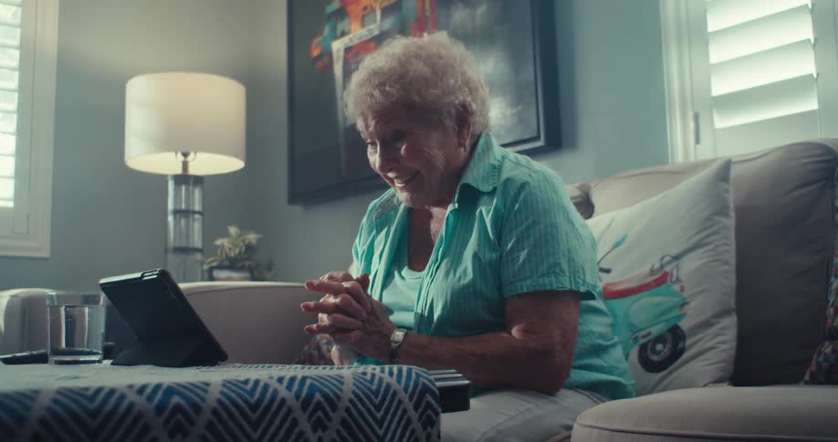 An elderly woman wearing a blue shirt is sitting on a sofa and is holding her hands in anxious tones looking at the screen of a tablet