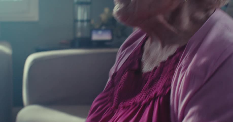 Elderly woman wearing a purple outfit sits on a couch with her hands interlocked together talking and smiling in her living room