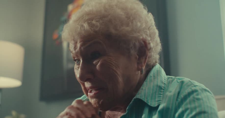 An elderly woman with short curly white hair her hands clasped looking upward with a sad expression in a softly lit room