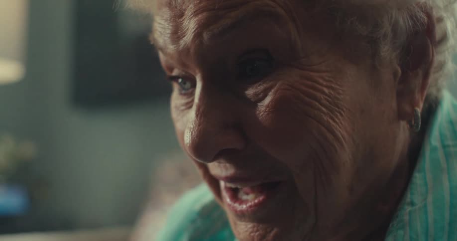 The face of an elderly woman who is wearing a blue shirt and she is talking while looking at something in front of her