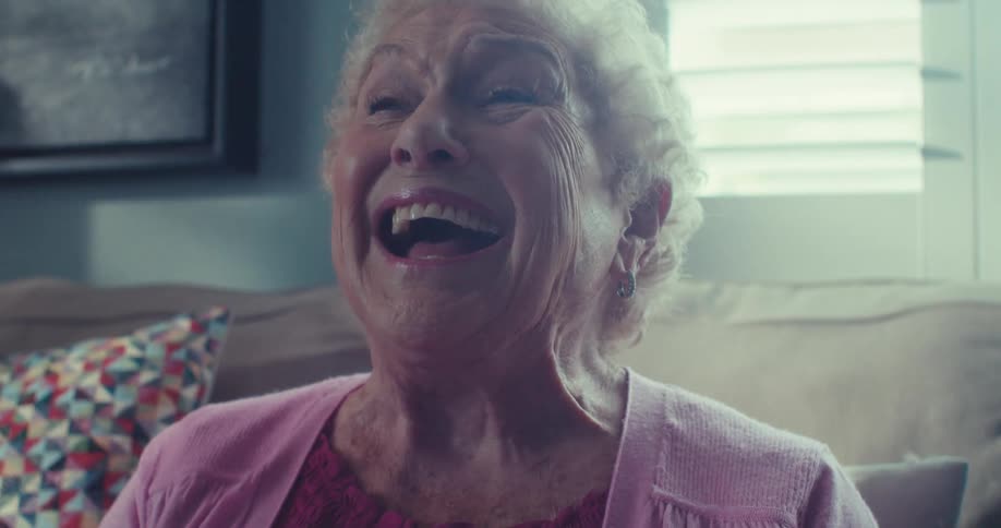 The face of an elderly woman in a pink shirt who is sitting on a sofa and is very happy while laughing and clapping her hands