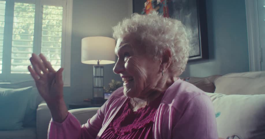 An older woman waves excitedly while smiling on a couch in a sunlit living room