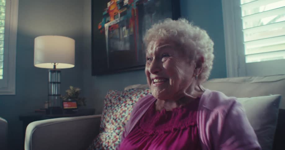 An excited elderly woman sitting on a couch waves her fists in the air as she smiles