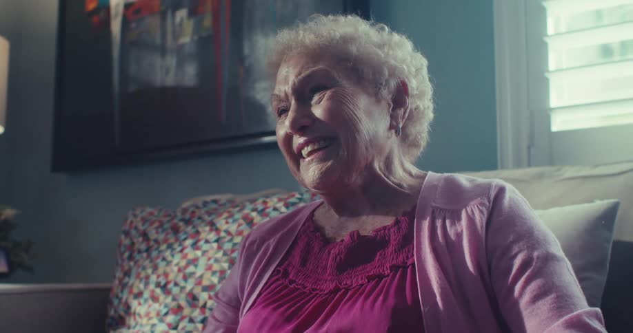 Elderly woman sits on the couch talking and laughing in her living room on a bright afternoon