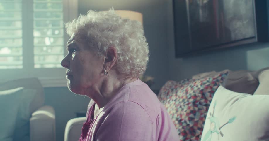 An elderly woman seated on her living room couch has a sad expression and shakes her head in disagreement