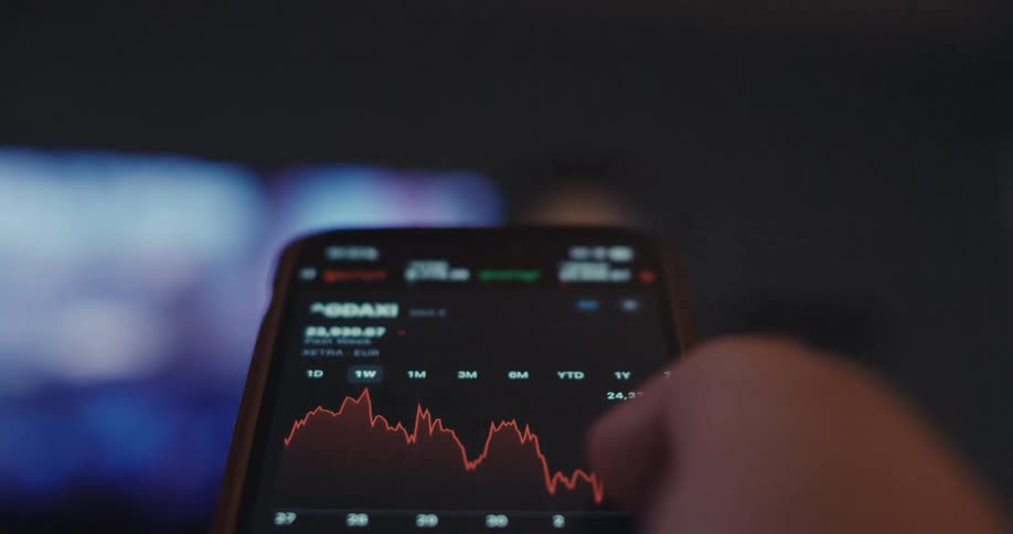 A closeup of a persons hand shows their thumb touching a smartphone screen displaying a financial line graph for the stock market