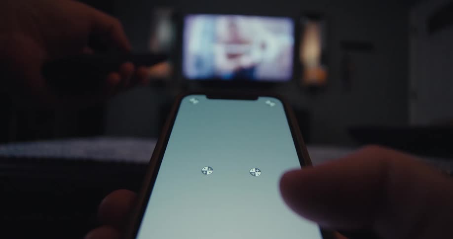 A person operates a mobile device and a tv remote control while sitting in a living room at night