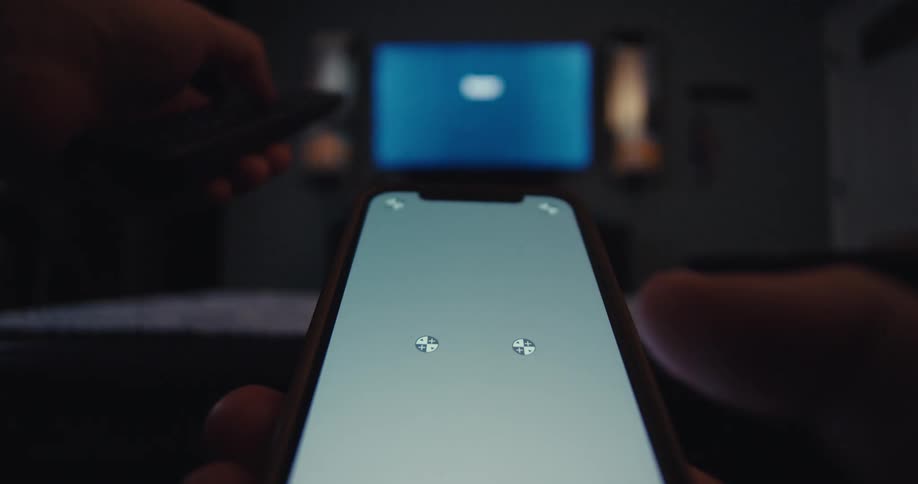 Man holds a smartphone while using a remote to change the television channel