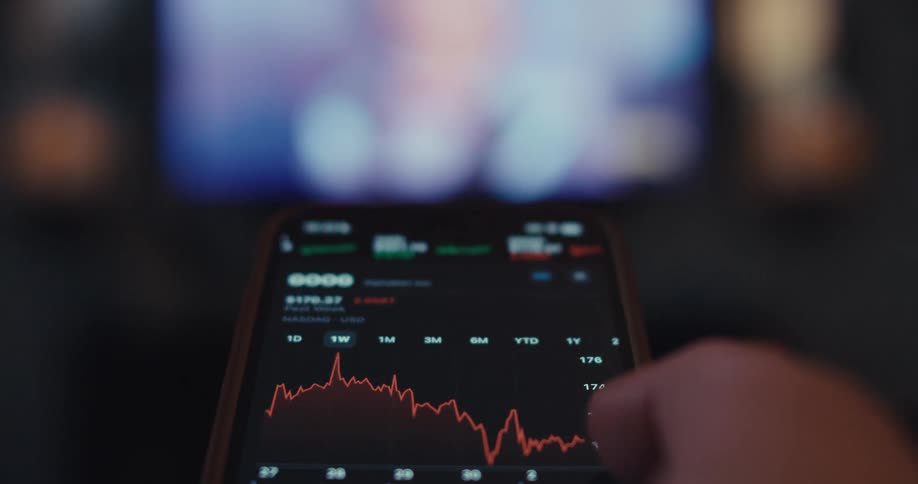 A person scrolls through various stock market charts on a smartphone app checking fluctuating prices with a television on in the background