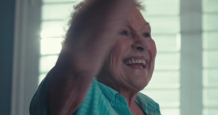 An elderly woman waves and happily dances while standing in a living room
