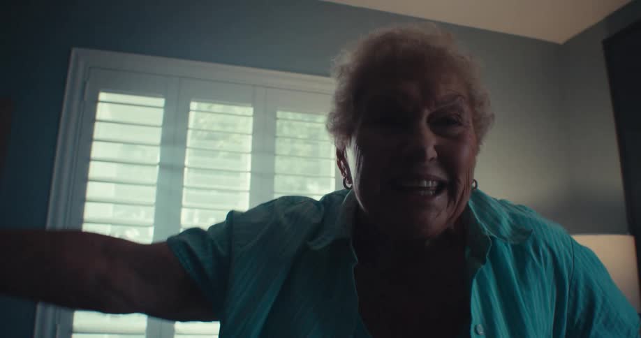 An elderly woman smiles joyfully while dancing inside a cozy sunlit living room