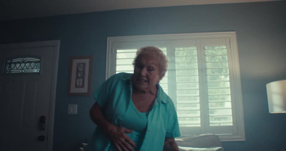 An elderly woman with short blonde hair energetically dances and sings in a living room her face illuminated by light from a window as the camera pulls back