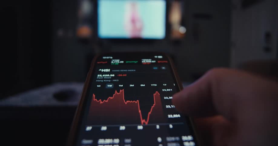 In a dimly lit room a finger scrolls through a fluctuating red stock market graph on a smartphone with a television glowing in the background