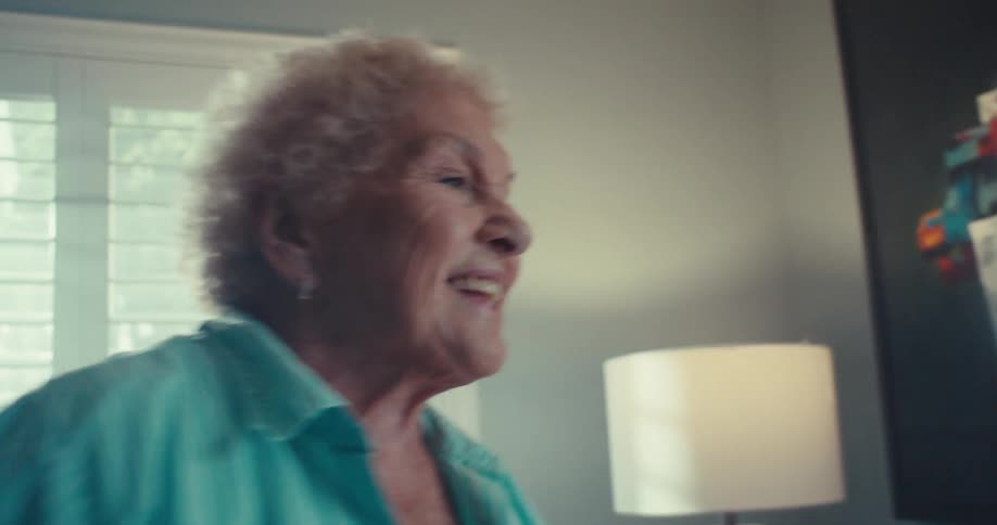 Elderly woman smiles and dances with her arms outstretched while standing in a living room