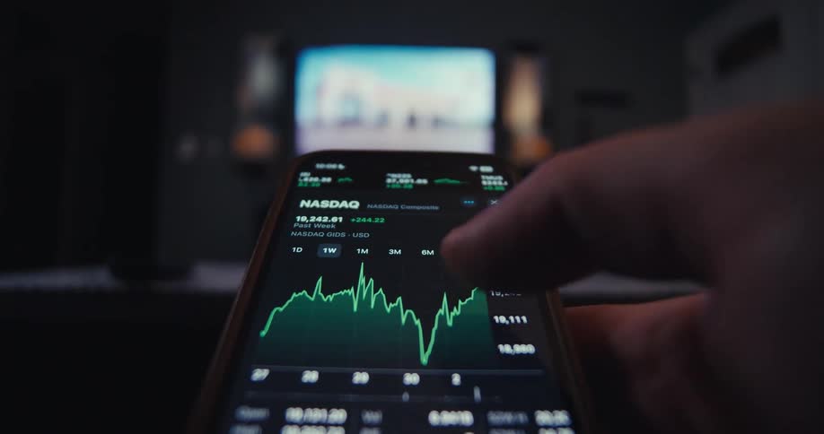 Person checks stock data on smartphone with blurred tv in background