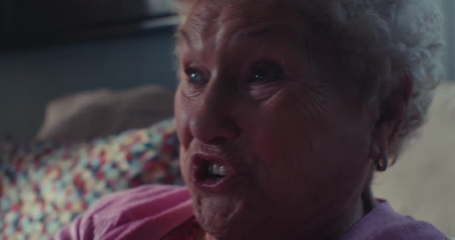 An elderly woman sits on a couch at midday holding a controller and gaming with excitement