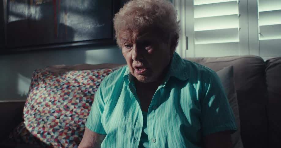 An elderly woman with short blonde hair sits on a couch in a dimly lit room speaking animatedly while looking at a tablet