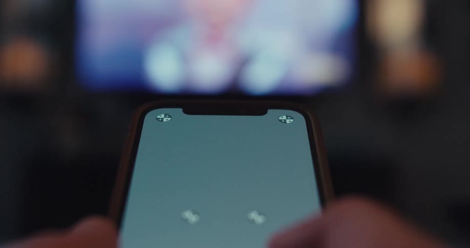 A firstperson perspective shows hands operating a smartphone with a grey replaceable screen while a blurred television displaying colorful content is visible in the dark background