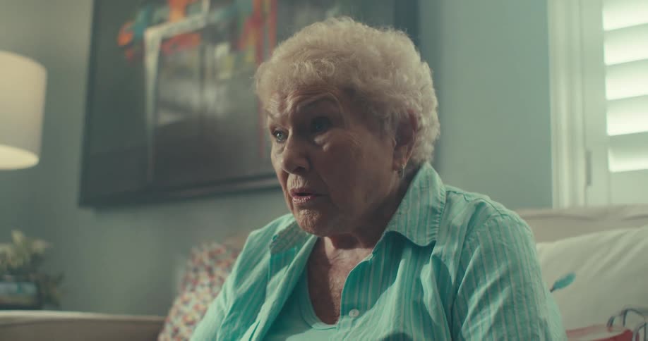 An elderly woman sits indoors with clasped hands looking emotional and tearful as she prays