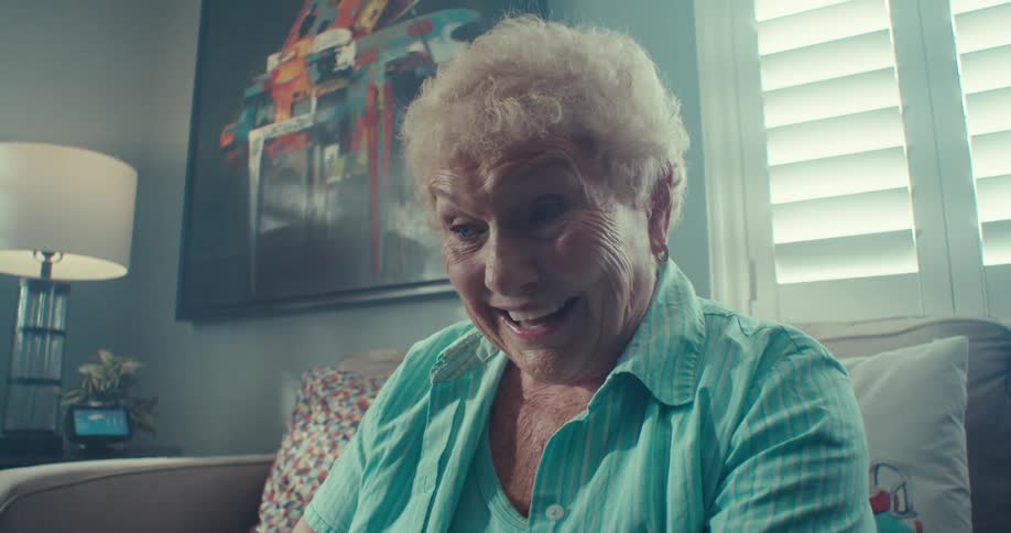 An elderly woman with a wide smile sits on a couch enthusiastically chatting on a laptop during a video call at home