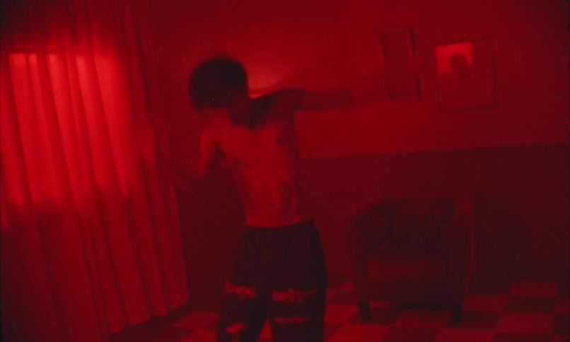 A shirtless man dances joyfully in a dimly lit redtinted room