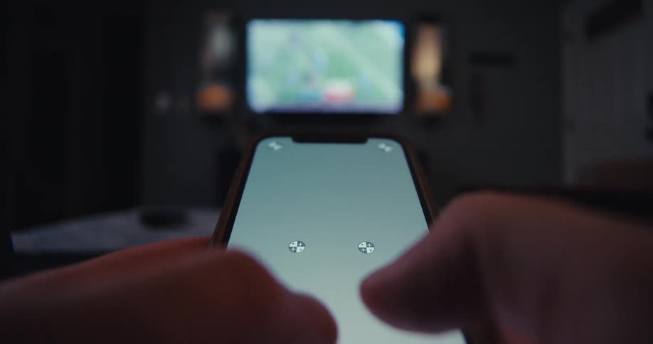 Person taps on smartphone screen while pointing it toward a blurred tv in a dark room