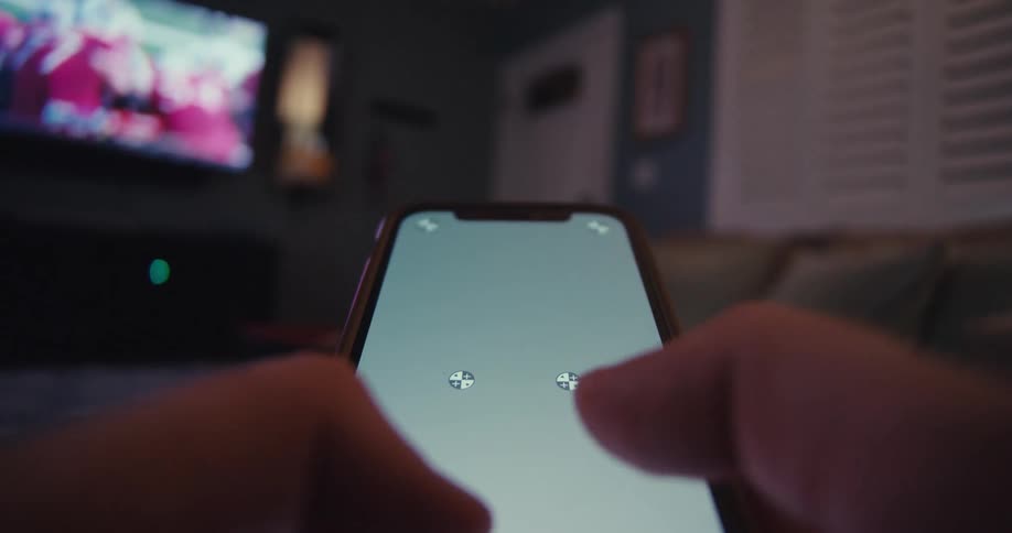 A person is holding a smartphone with a blank screen displaying two small crosshair icons while a television is visible in the background
