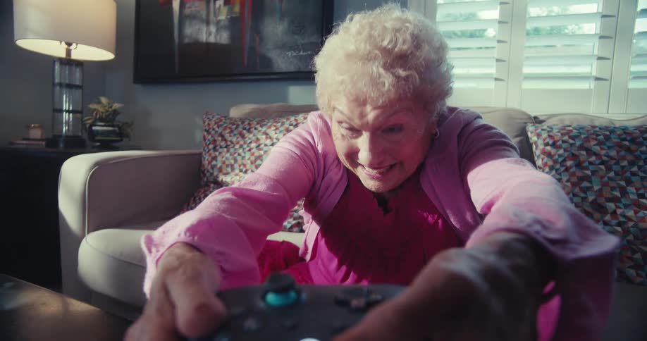 A cheerful elderly woman with short curly white hair and a pink shirt playing video games looking intensely at a screen laughing