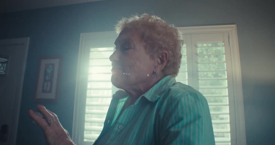An elderly woman in a blue shirt is happily dancing indoors near a window