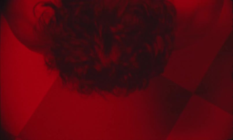 Man lying on a check board patterned floor in a red lit room moves his head backward and forward